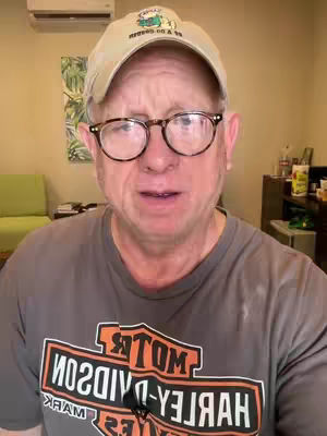 A middle-aged man with glasses and a baseball cap looks directly at the camera. He is wearing a t-shirt with a motorcycle logo. The background is a simple interior setting.