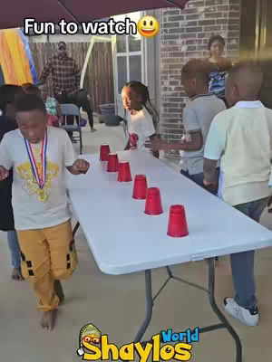 A group of children are gathered in an outdoor setting. One child is running toward the camera while wearing a medal. A white table lined with red cups is set up in front of the children. Other adults and children are visible in the background.