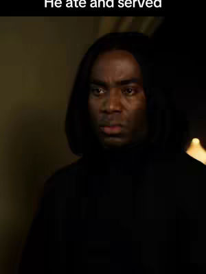 A medium shot features a man with a dark complexion, wearing a black hooded garment. The man is looking towards the left with a contemplative expression, showing slight furrowing of the brows. The background is dimly lit, with soft shadows suggesting an indoor setting.