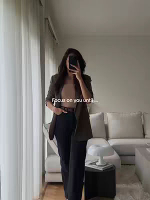 A woman stands in a well-lit living room, taking a selfie. She is dressed in a professional outfit, dark trousers, and a blazer, holding a phone in front of her face. The background shows a neutral-toned sofa, window, and curtains, indicating a modern and stylish interior.
