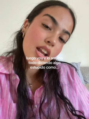 The image features a young woman in a close-up, angled shot. She has long, dark hair, styled away from her face, and visible makeup. She wears a pink, striped button-down shirt and a small necklace. The background is blurred, suggesting an indoor setting.