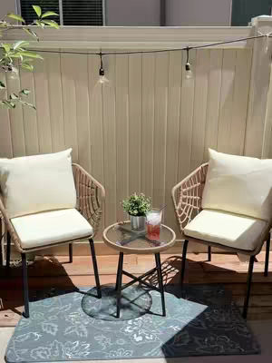 The image shows an outdoor patio scene. Two wicker chairs with beige cushions face each other, with a small round table between them. A plant and a glass of drink sit on the table. The background is a wooden fence with string lights hanging across. There is a patterned rug on the ground.