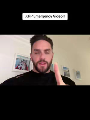 A man with a beard and dark hair is directly facing the camera, pointing upwards with his right index finger. Above him, the text 'XRP Emergency Video!!' is displayed. Behind him are white walls with paintings displayed. The image is framed vertically, with a black background filling the bottom portion.