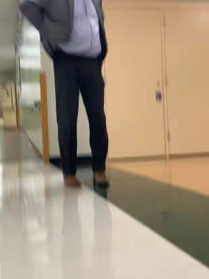 A low-angle shot in a brightly lit hallway with a polished floor. A person wearing a suit jacket over a button-down shirt and dark trousers is standing with their hands on their hips.