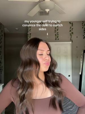 The image displays a woman with long, wavy brown hair, wearing a brown long-sleeved top. Her expression is a slight smirk with closed eyes. Above her, a ceiling fan is visible. The text overlay reads, 'my younger self trying to convince the devil to switch sides'.