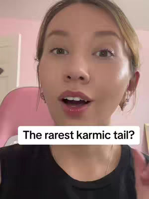 A close-up shot of a woman with light skin and light brown hair, looking directly at the camera. She has a slight smile, and her mouth is open as if speaking. The background is a blurred interior with pink elements.