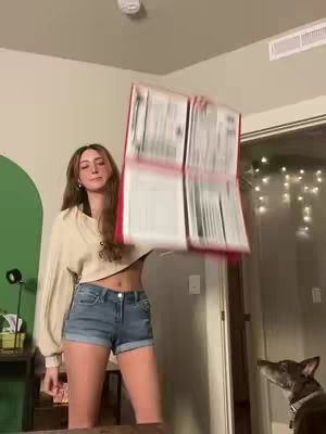 A woman in a cropped beige sweater and denim shorts is holding up a large, open notebook with red binding. She is smiling and looking at the camera. In the background, there is a dog looking up at the notebook.