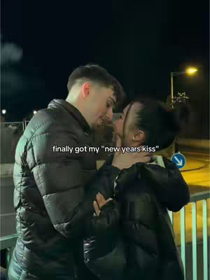 A couple is embracing and about to kiss outdoors at night. The man, on the left, is turned slightly towards the woman, who's on the right. They're wearing dark, puffy jackets. The background is blurred, featuring some lights and a blue directional arrow.