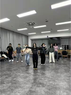 A medium shot of a group of people in a large room. Seven people are present, standing on a gray floor. The person in the center, a female, stands with her arms outstretched, and is wearing a black jacket, black pants, and light-brown boots. Several people around her are also putting their arms out with her. The background consists of a white ceiling adorned with lights, and white curtains with some tables. There s a vent in the ceiling.