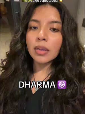 A woman with long, dark wavy hair looks directly at the viewer. She is wearing a dark v-neck top. Text overlays the image, with the word “DHARMA” in white, bold font, and a purple wheel-shaped symbol. Some indoor background is visible.