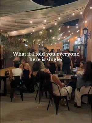 An outdoor scene at night with a patio. Strings of lights are hanging above a crowd of people in a bar or restaurant setting. The main focus is on the people, who are sitting and standing, engaged in conversation. The lighting is warm, creating a lively and inviting ambiance.