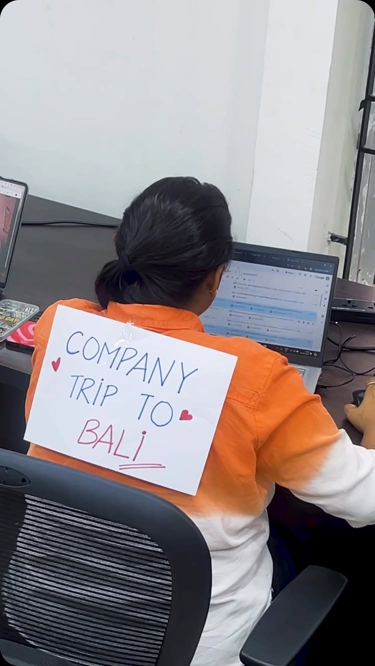 A woman is seated at a desk in an office, viewed from behind. She faces a laptop displaying a screen, seemingly in the midst of work. Affixed to her back is a white sheet of paper bearing the handwritten message "COMPANY TRIP TO BALI," adorned with small heart symbols. The scene is brightly lit, and the composition is focused on the work environment with the woman as the central subject.