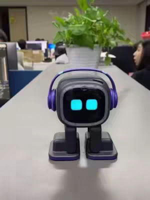 The cover image features a small, dark gray robot with a cubical head and large blue eyes, wearing purple headphones. It is positioned on a slightly reflective white surface with a plant in a pot behind it. The background shows parts of an office setting with desks and people.