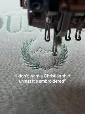 The image shows a close-up of an embroidery machine in operation, creating a design on light-colored fabric. The machine's needle is positioned over a design of leaves and mountains. Text appears in a clean font at the bottom of the image, stating a preference for embroidered Christian shirts.