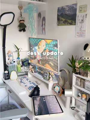 A well-lit desk setup with various anime-themed items. A computer monitor displays colorful animation alongside assorted figurines, plants, and desk organizers, creating a vibrant workspace aesthetic.