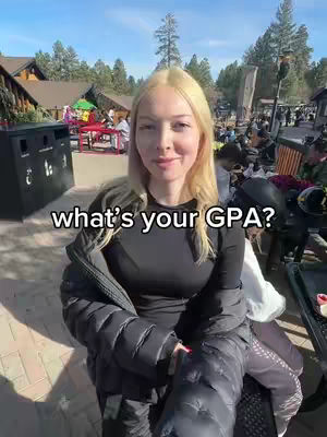 A young woman with blonde hair sits outdoors, facing the camera with a slight smile. She wears a black top and a black jacket, and the background shows trees, buildings, and other people, suggesting an outdoor event or location. Text overlay reads "what's your GPA?"