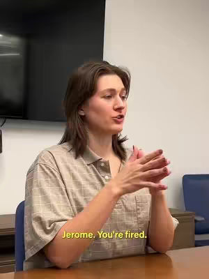 A woman with medium-length brown hair sits at a table and gestures with her hands as she speaks. The scene has a simple background with a dark panel on the wall and a table.