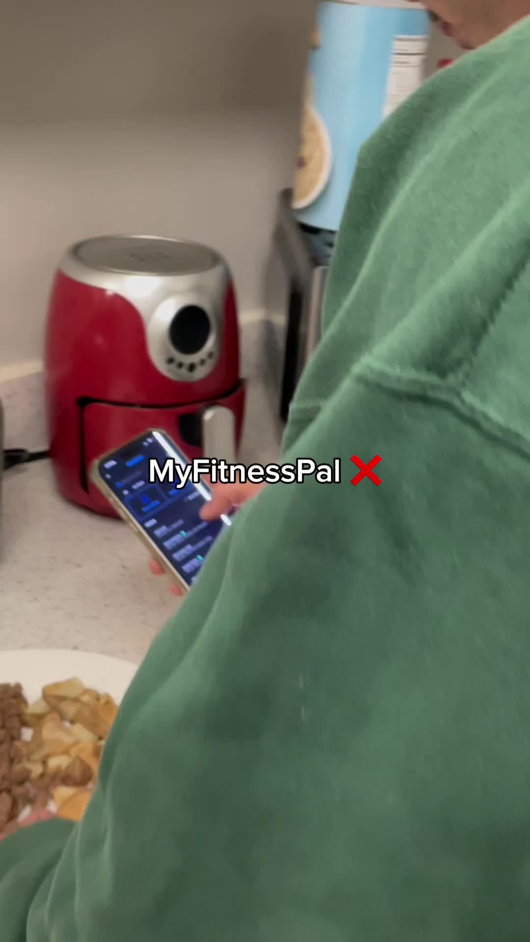 A person, seen from the chest up, is holding a phone showing an app interface with data displays. Overlayed text spells out 'MyFitnessPal' with a red 'X' next to it. The background is a kitchen with a red air fryer and other cooking supplies.