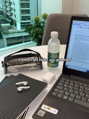 A well-lit shot featuring a desk setup. A laptop is open, displaying a document, accompanied by a bottle of Coco Legal water, two notebooks, a pencil case, and wireless earbuds. An office style window and chair complete the scene.