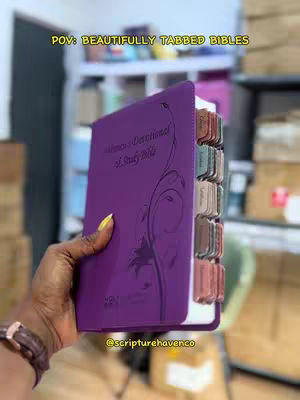 A person's hand holds open a purple Bible with tabbed pages, with a background of shelves. The hand is dark-skinned with a gold watch and yellow nail polish. The Bible has intricate swirls embossed on the front. The tabs on the side have different colors.