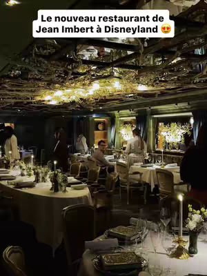 An interior shot of a restaurant with an ornate ceiling and tables set for dining. The ceiling is covered with foliage, and the lighting is dim. Tables are set with white tablecloths, candles, and flowers. People are seated, suggesting a busy and elegant dining environment.