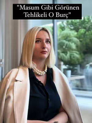 A woman with blonde hair wearing a black shirt and cream-colored jacket, a pearl necklace, posing indoors with a blurred background of large windows and greenery.