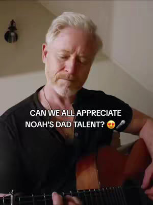 A middle-aged man with light hair and a beard plays an acoustic guitar in what appears to be a warmly lit room. The text overlay invites viewers to appreciate his talent.