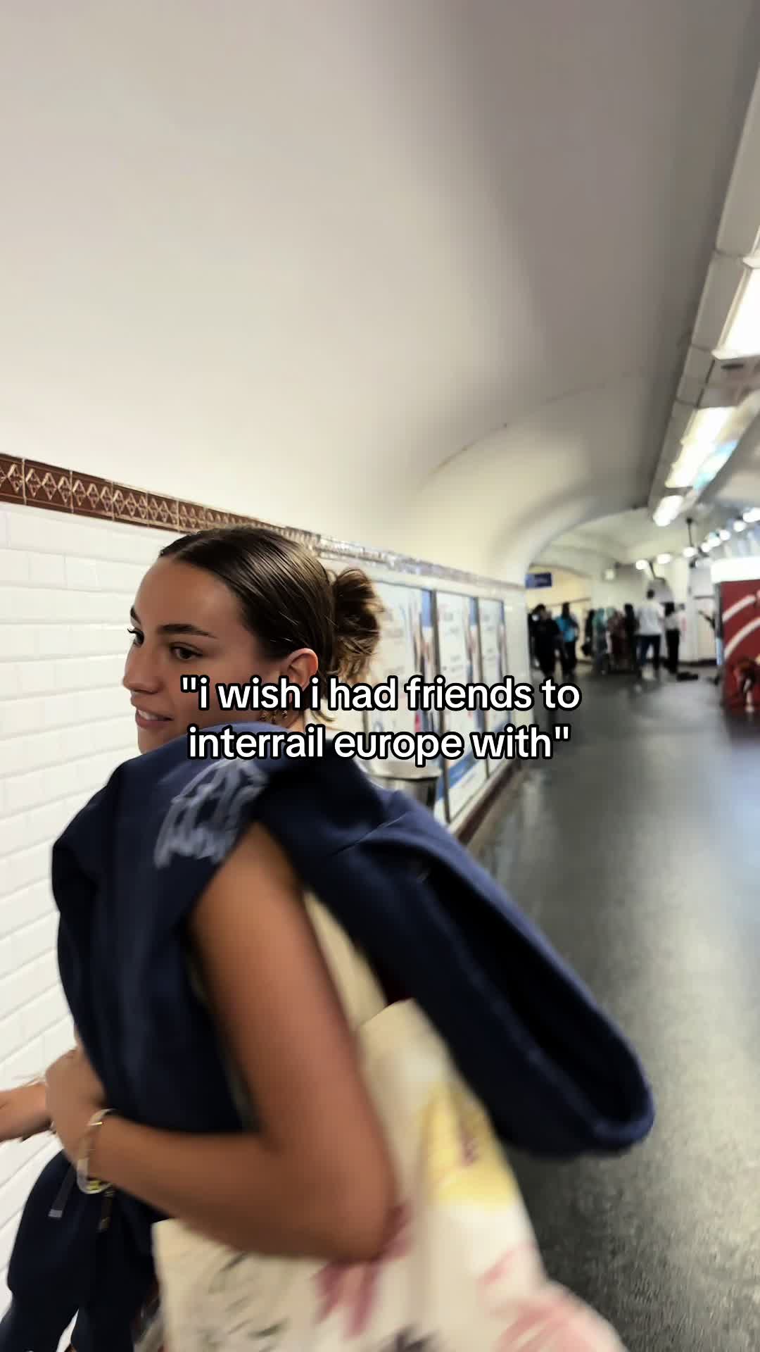 A woman walks through a subway station, looking to her left. The background includes a white-tiled wall with a decorative ledge and advertisements, suggesting an urban environment. The image has a slightly blurred effect.