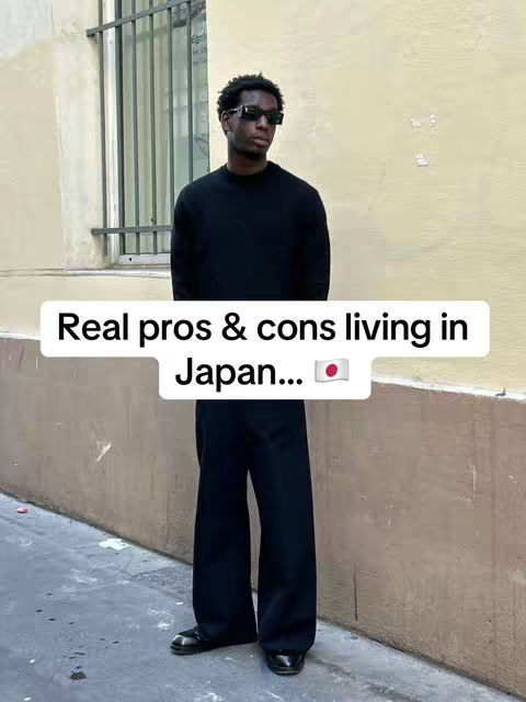 A person in a black sweater and pants stands against a yellow wall. A white text box overlaid on the image reads 'Real pros & cons living in Japan...' with the Japan flag.