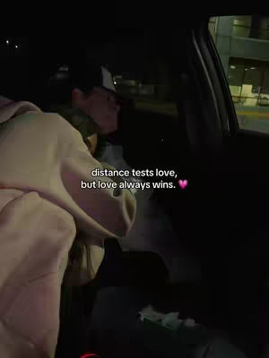 The image shows a person in a car at night. The main subject is in the foreground, wrapped in a pinkish hoodie. The text overlayed on the image says, "distance tests love, but love always wins." There's a small heart emoji. The car interior and darkness imply the video is being filmed at dusk.
