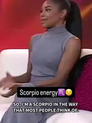A woman with dark hair is seated on a white couch in a brightly lit setting. She is wearing a sleeveless gray dress and gesturing with her hands as she talks. A black bar with white text overlays part of the scene.