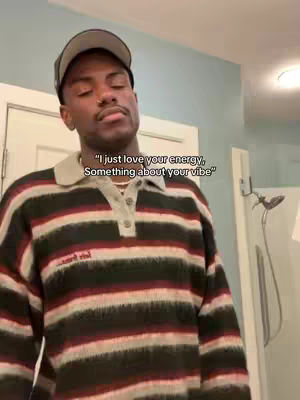 The image shows a young man in a bathroom, looking up and to the side with a thoughtful expression. He's wearing a striped sweater, a gray baseball cap, and a silver necklace. Text overlayed in the image says, "I just love your energy, Something about your vibe". He appears to be in a lit bathroom with a partial view of a shower.