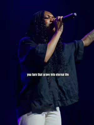 A stage performance shot with a person holding a microphone and singing. The person is wearing a black top and white pants, with a tattoo visible on their arm. Overlayed text reads, 'you turn that grave into eternal life'. The background is dark, with blue and purple lighting.