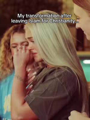 A medium shot shows three women in a warm-toned setting. The central figure, with long blonde hair, is the main focus, partially blocking the face of the woman next to her. The main woman is holding the other's arm and appears emotional. The background consists of blurred colors and out-of-focus forms, suggesting an interior setting.