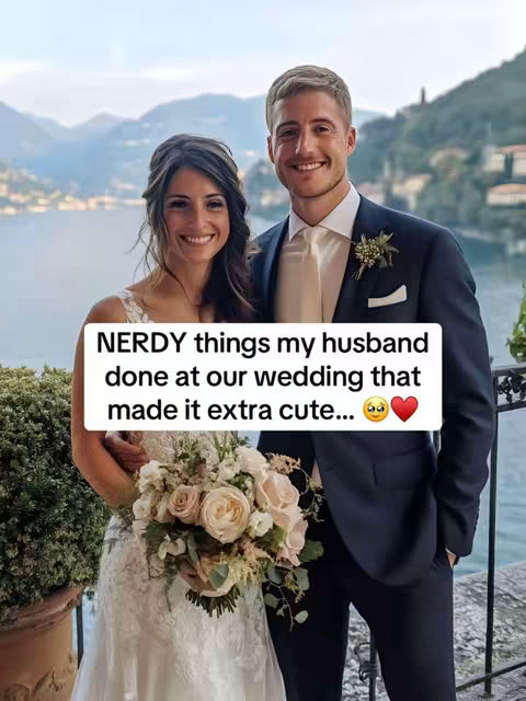 A couple stands closely together, facing the camera. The woman is in a white wedding dress and holding a bouquet of flowers. The man is wearing a navy suit and smiling. In the background are mountains and a body of water.