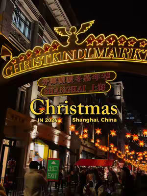 A night scene at a Christmas market, framed with a lit-up sign that reads "CHRISTKINDLMARKT" and the Chinese characters. Below, the text "Christmas IN 2024 Shanghai, China" is overlaid. The image captures a bustling street scene with many people, vibrant lighting, and decorative stars.