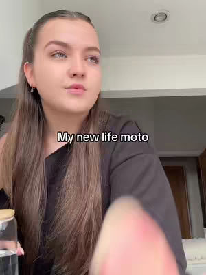 A woman with long brown hair, wearing a black shirt, sits indoors. The top of a clear glass is visible in her left hand. Text 'My new life moto' appears as an overlay.