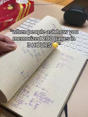 A person's hand holding open a notebook filled with handwritten notes and mathematical equations. The background includes a white keyboard, a black mouse and a red box of treats.
