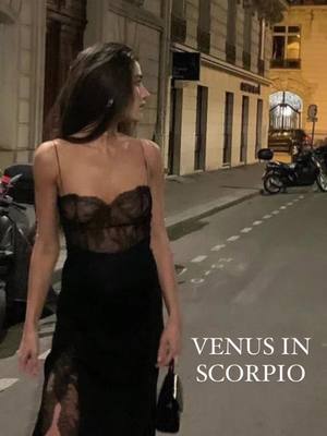 A woman in a black dress with spaghetti straps and lace detailing is shown walking on a city street at night. The image is softly lit, with the background showing buildings and street lights. Text overlays the image, mentioning "Venus in Scorpio."