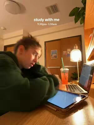 A young woman is studying at a desk in her room. She is wearing a green hoodie and is leaning over a laptop and a tablet. There is a lamp, a drink, and a plant on the desk as well. The background is a light-colored wall with a door that has some posters on it.