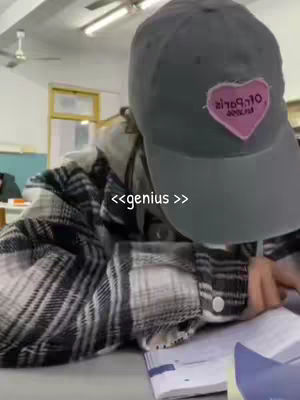 A person wearing a plaid shirt and a baseball cap is leaning over a book. The cap is gray with a pink heart-shaped logo. The person is in a room with fluorescent lights.