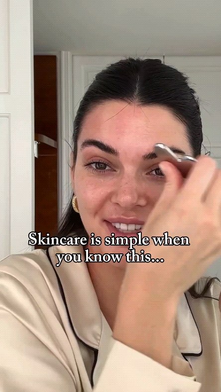 A woman with dark hair is applying something to her eyebrow. She is wearing a cream-colored top and gold earrings. Text overlayed on the image is 'Skincare is simple when you know this...'