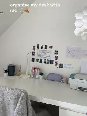 A well-organized study desk with a white surface is the primary focus. The background includes a white wall with a sloped ceiling, and a geometric light fixture. On the desk are stationery items, a desk lamp, a small potted plant, and a printer. There is a soft blanket draped over a chair in the lower left corner.