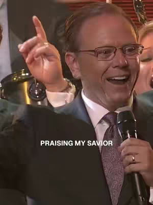 A man in a dark suit with a patterned tie, glasses, and short hair is holding a microphone and speaking. The image has a warm tone and is well-lit, conveying a sense of positivity and reverence.