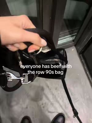 The cover image shows a close-up shot of a black shoulder bag being unzipped. The hand is holding the bag with the zipper open. The background has a hint of a window or door. The text overlay reads: "everyone has beef with the row 90s bag".