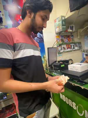 A man stands in a store looking down at what appears to be a stack of bills held in his hands. The store has a green counter with the words 'NOORS' visible. Shelves in the background contain various items. The man wears a black, white, and red striped shirt.