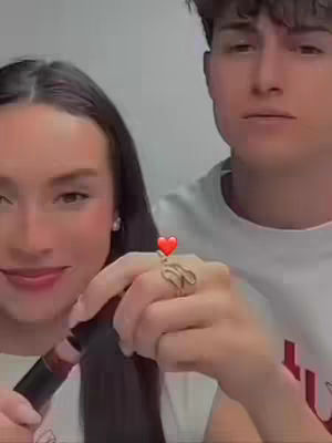 The image presents a couple, with the woman in the foreground applying lipstick. She is facing the camera and smiling while glancing downwards towards her hand that holds the lipstick. In the background, the man also smiling, holds the woman's hand with a ring on it. Colors are warm, with natural skin tones predominating and the background is a neutral white. The focus is on the couple's affectionate interaction, communicating a sense of love and connection.