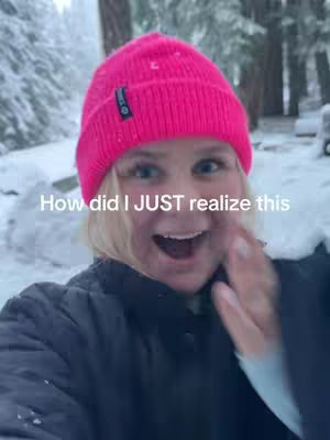 A woman with blonde hair wearing a bright pink beanie smiles at the camera with snow falling around her. The background shows snow-covered trees.