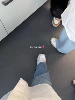 A low-angle shot captures three individuals' legs and feet with a grey floor. The person in the foreground has white sneakers and blue jeans, while others have different shoes and black pants. The word 'medicine.' is written in the center of the frame, near one of the feet, with a small heart emoji.