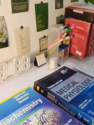 A well-lit image displays a desk setup with study materials. Several textbooks, including titles like 'Textbook of Medical Physiology' and 'Biochemistry,' are visible, along with a digital clock, sticky notes, pens, and a small calendar. The composition suggests a study environment, likely for medical students.